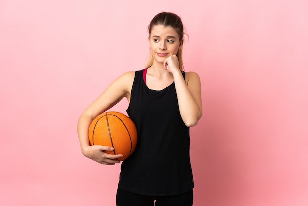 Young blonde woman playing basketball isolated on pink wall having doubts and thinking