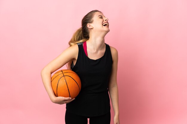 Young blonde woman playing basketball isolated on pink wall laughing in lateral position