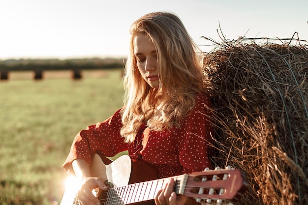 Young blonde woman plays guitar near a haystack in a field in the sunset