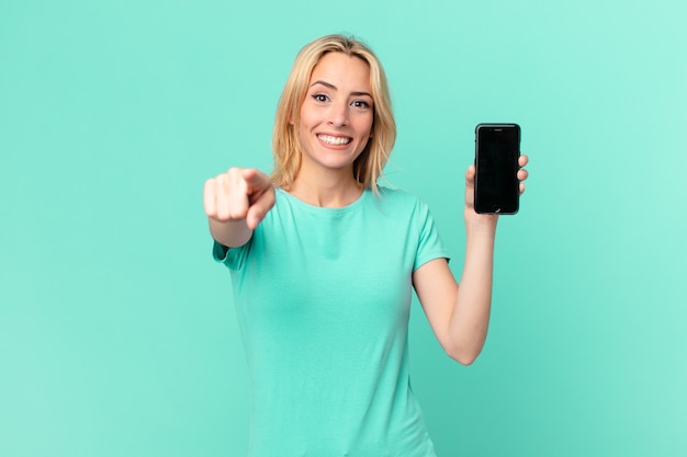 Young blonde woman pointing at camera choosing you and holding a smart phone