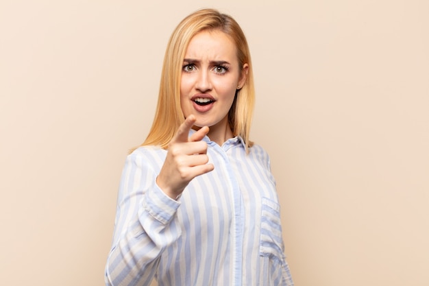 Young blonde woman pointing at camera with an angry aggressive expression looking like a furious, crazy boss