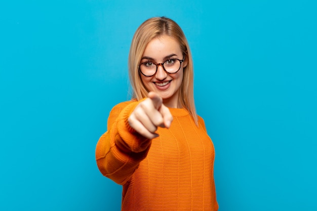 Young blonde woman pointing at front with a satisfied, confident, friendly smile, choosing you