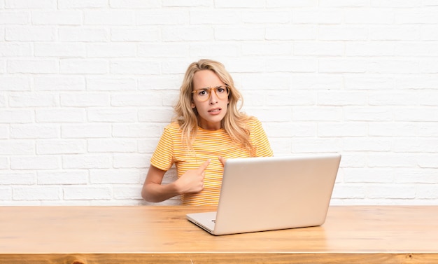 Young blonde woman pointing to self with a confused and quizzical look, shocked and surprised to be chosen using a laptop