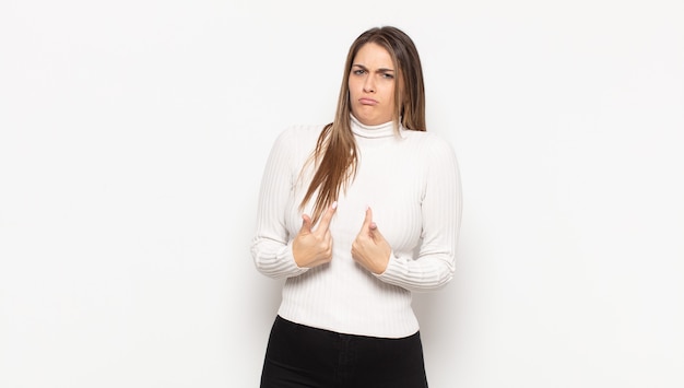 Young blonde woman pointing to self with a confused and quizzical look, shocked and surprised to be chosen