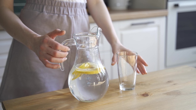 A young blonde woman pours lemon water from a decanter into a glass. Refreshing water with lemon slices. 4K UHD