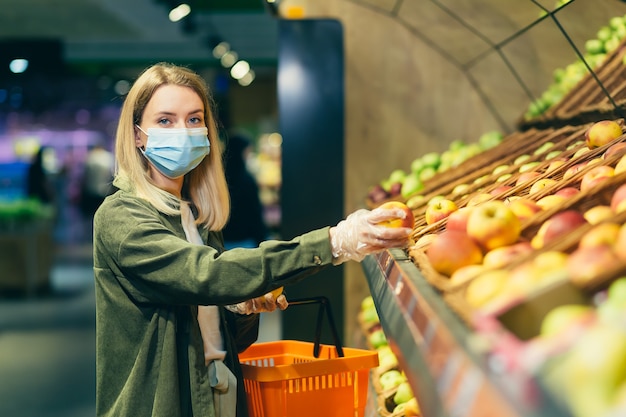 Young blonde woman in a protective face medical mask picks chooses apple fruits vegetables on the counter in supermarket. Female shopping in market standing near department store with basket in hands