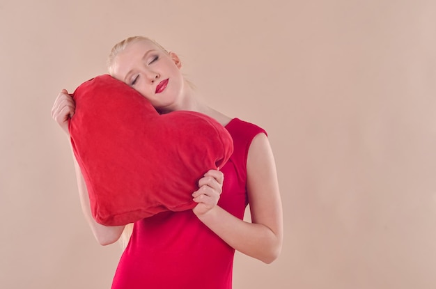 Young blonde woman in a red dress holds a red heart in her hands on a beige background