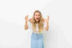 Photo young blonde woman screaming in panic or anger, shocked, terrified or furious, with hands next to head against white wall