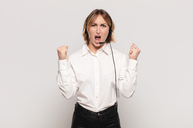 Young blonde woman shouting aggressively with an angry expression or with fists clenched celebrating success