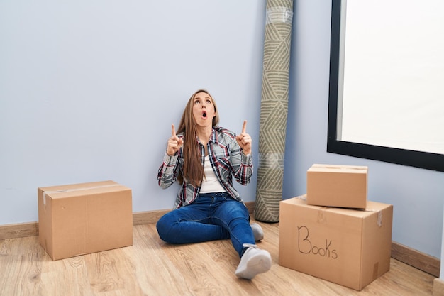 Young blonde woman sitting on the floor moving to a new home amazed and surprised looking up and pointing with fingers and raised arms