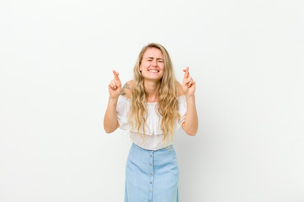 Young blonde woman smiling and anxiously crossing both fingers, feeling worried and wishing or hoping for good luck against white wall