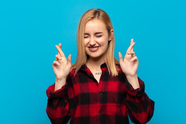 Young blonde woman smiling and anxiously crossing both fingers, feeling worried and wishing or hoping for good luck