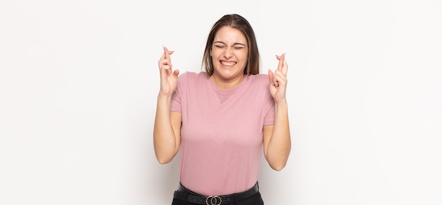 Young blonde woman smiling and anxiously crossing both fingers, feeling worried and wishing or hoping for good luck