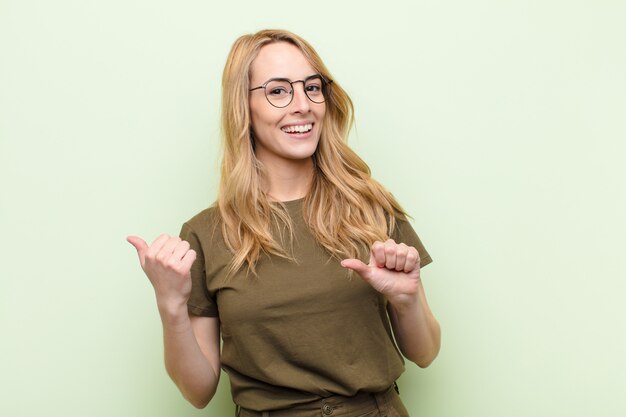 Young blonde woman smiling cheerfully and casually pointing to copy space on the side, feeling happy and satisfied on flat color wall