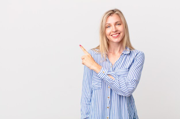 young blonde woman smiling cheerfully, feeling happy and pointing to the side