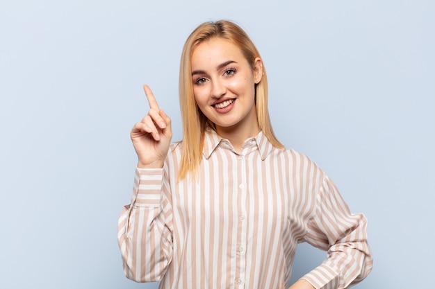 Young blonde woman smiling cheerfully and happily, pointing upwards with one hand to copy space