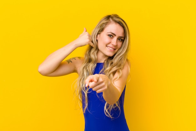 Young blonde woman smiling cheerfully and pointing to camera while making a call you later gesture, talking on phone against yellow wall