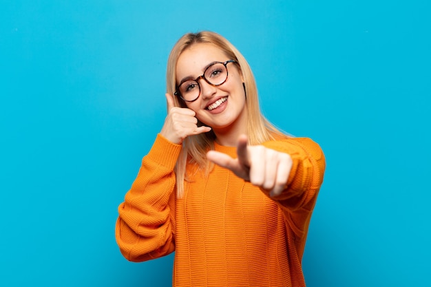 Young blonde woman smiling cheerfully and pointing to camera while making a call you later gesture, talking on phone