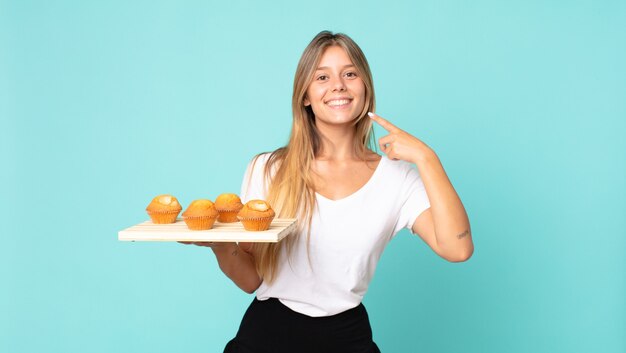 Young blonde woman smiling confidently pointing to own broad smile and holding a muffins troy