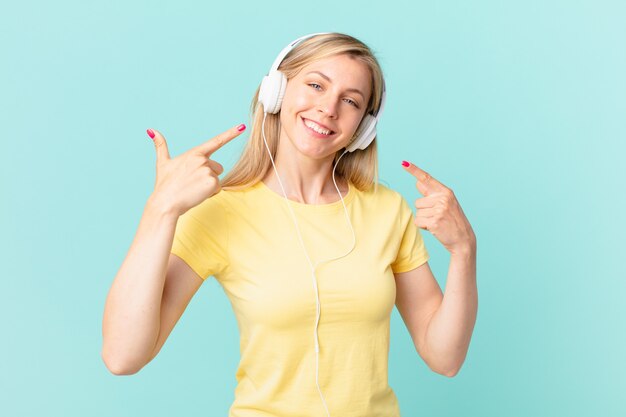 Young blonde woman smiling confidently pointing to own broad smile and listening music.