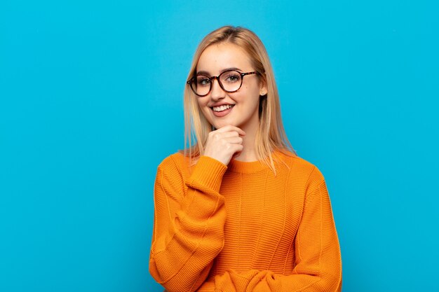 Giovane donna bionda sorridente, godersi la vita, sentirsi felice, amichevole, soddisfatta e spensierata con la mano sul mento