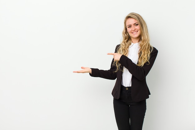 Photo young blonde woman smiling, feeling happy, carefree and satisfied, pointing to concept or idea on copy space on the side against white wall
