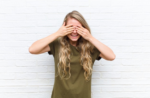 Photo young blonde woman smiling and feeling happy, covering eyes with both hands and waiting for unbelievable surprise against brick wall