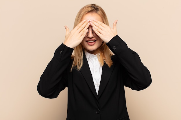 Young blonde woman smiling and feeling happy, covering eyes with both hands and waiting for unbelievable surprise