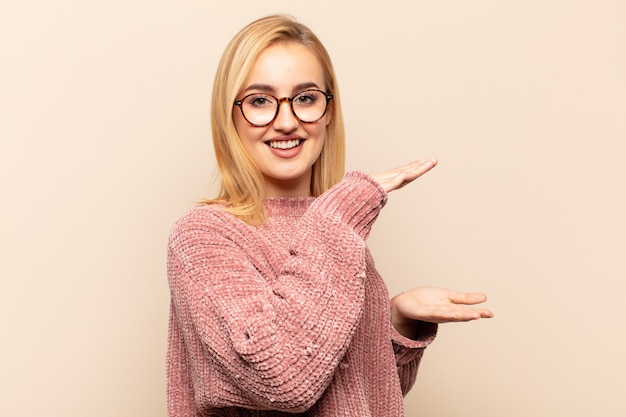 Young blonde woman smiling, feeling happy, positive and satisfied, holding or showing object or concept on copy space