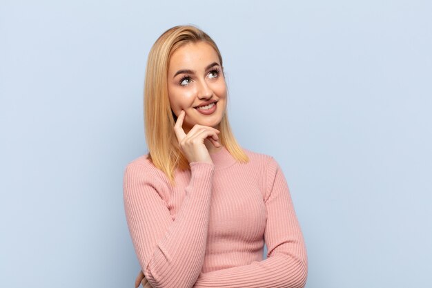 Young blonde woman smiling happily and daydreaming or doubting, looking to the side