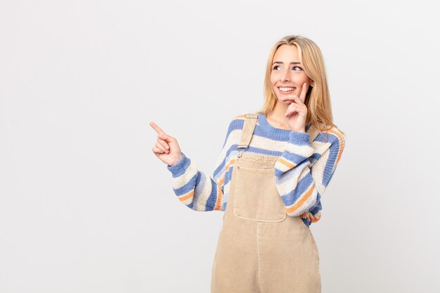 Young blonde woman smiling happily and daydreaming or doubting