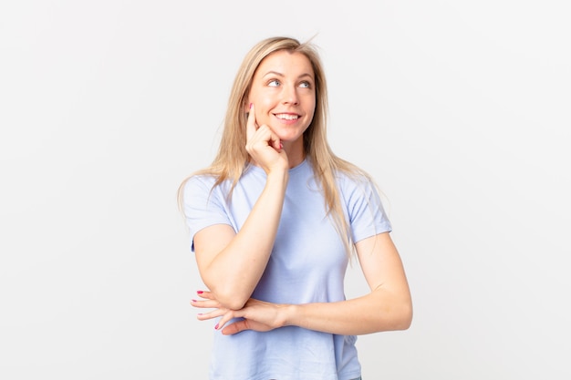 Young blonde woman smiling happily and daydreaming or doubting