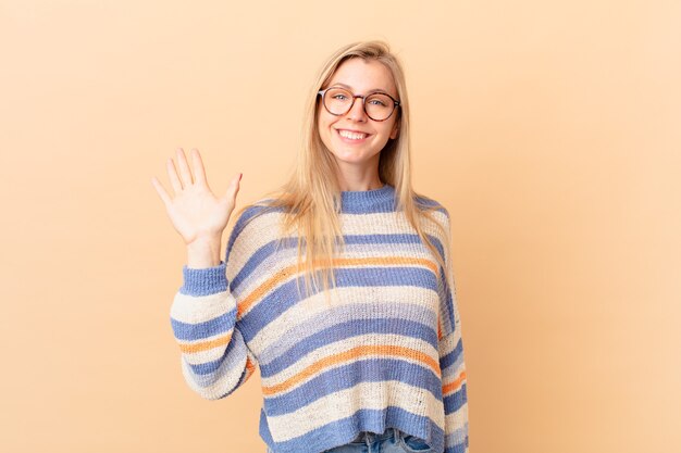 Young blonde woman smiling happily, waving hand, welcoming and greeting you