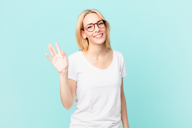 Young blonde woman smiling happily, waving hand, welcoming and greeting you