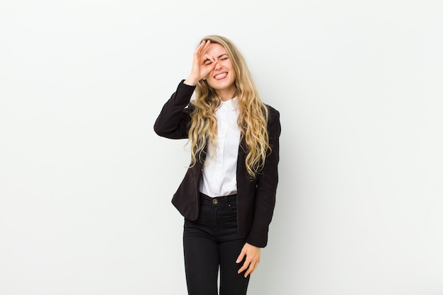 Young blonde woman smiling happily with funny face, joking and looking through peephole, spying on secrets against white wall