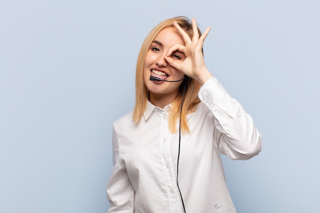 Young blonde woman smiling happily with funny face, joking and looking through peephole, spying on secrets