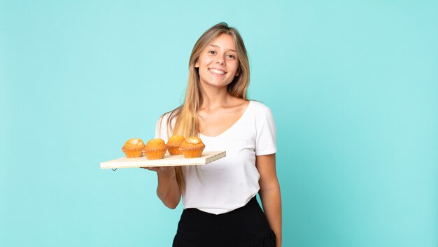 Young blonde woman smiling happily with a hand on hip and confident and holding a muffins troy