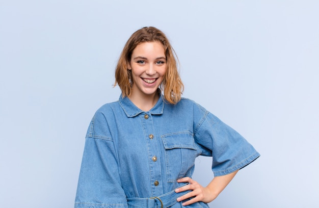 Young blonde woman smiling happily with a hand on hip and confident, positive, proud and friendly attitude against flat color wall