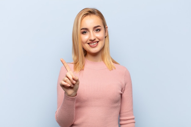 Young blonde woman smiling and looking friendly, showing number one or first with hand forward, counting down