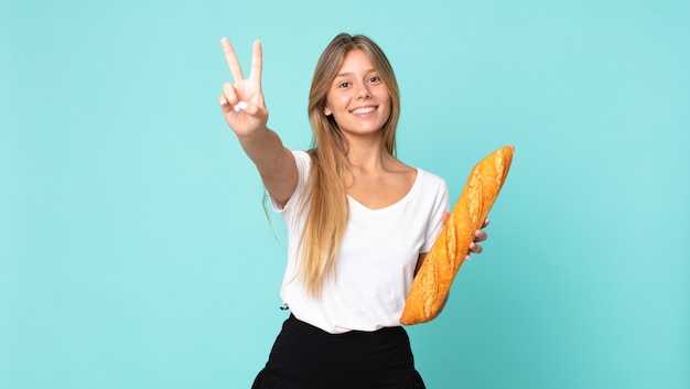 Young blonde woman smiling and looking happy, gesturing victory or peace and holding a bread baguette