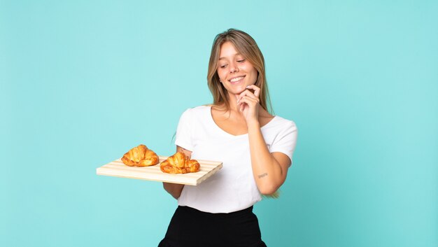 Young blonde woman smiling with a happy, confident expression with hand on chin and holding a croissant tray