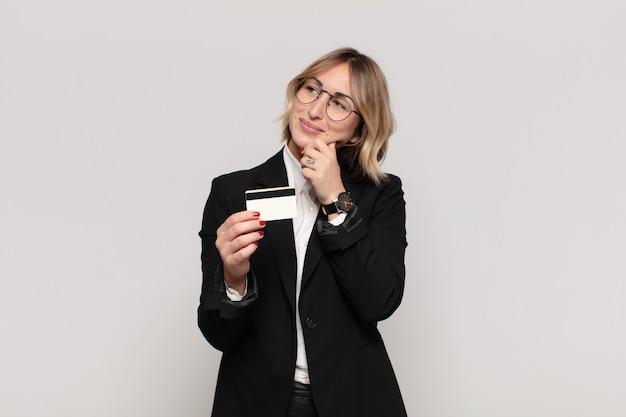 Young blonde woman smiling with a happy, confident expression with hand on chin, wondering and looking to the side