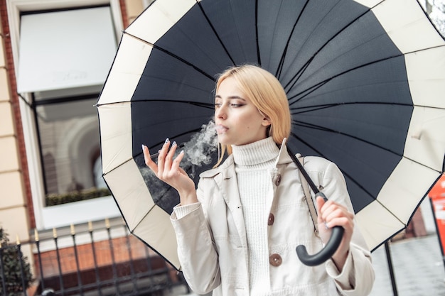 Young blonde woman smokes electronic cigarette under umbrella in the city