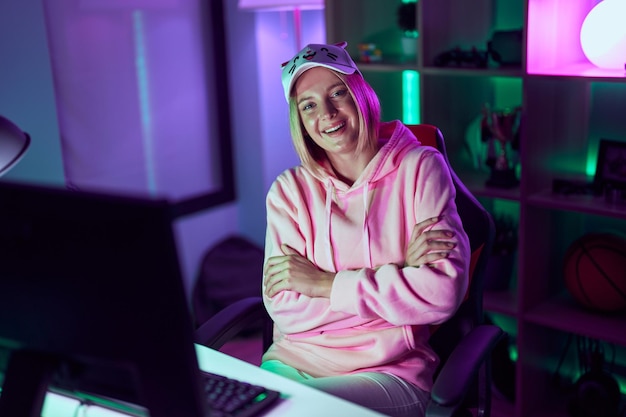 Young blonde woman streamer smiling confident sitting with arms crossed gesture at gaming room