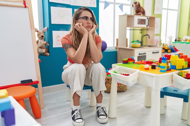 Young blonde woman teacher sitting on chair with serious expression at kindergarten