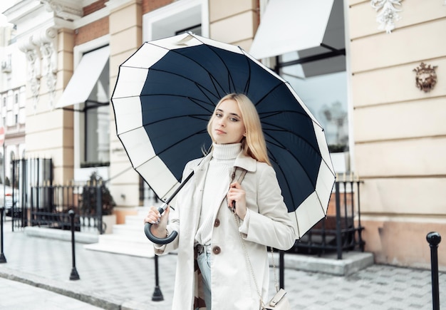 Giovane donna bionda in trench con ombrello in città