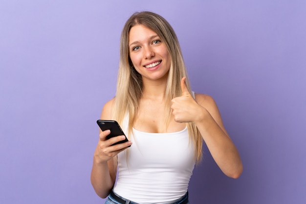 Young blonde woman using mobile phone isolated on purple wall giving a thumbs up gesture