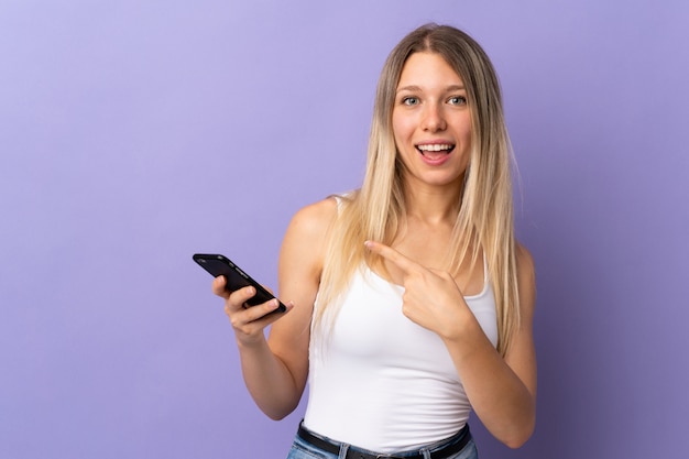 Young blonde woman using mobile phone isolated on purple wall pointing finger to the side