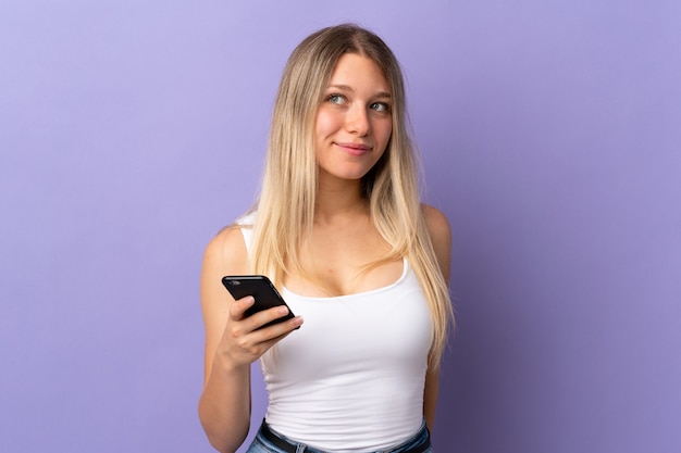 Young blonde woman using mobile phone isolated on purple wall standing and looking to the side