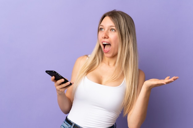 Young blonde woman using mobile phone isolated on purple wall with surprise facial expression